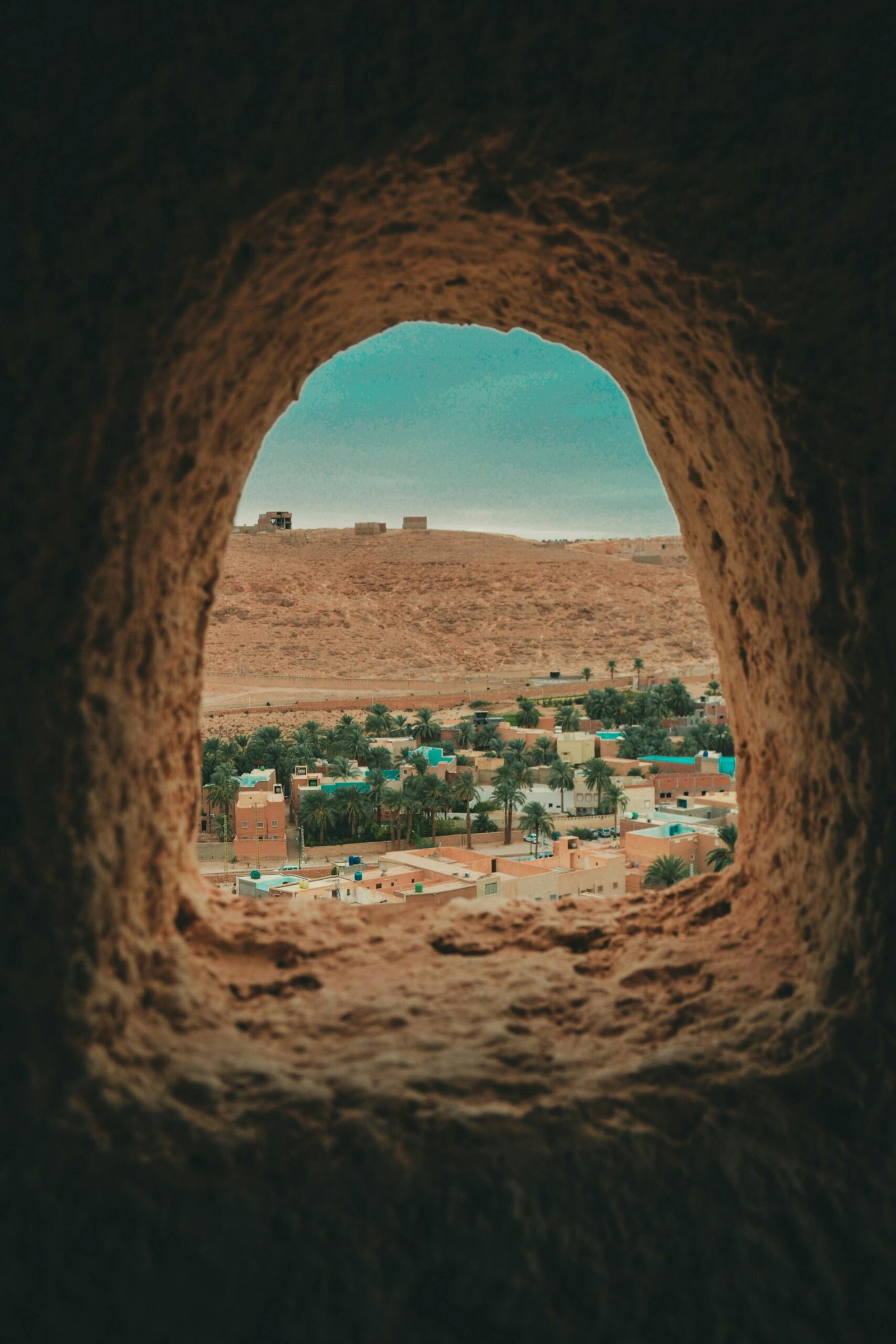 View through a cave opening revealing a desert town with palm trees and hills.